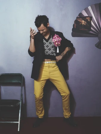 Harry Styles wearing yellow trousers, a cropped geometric vest, and a black blazer with a pink corsage posing against a lilac wall.