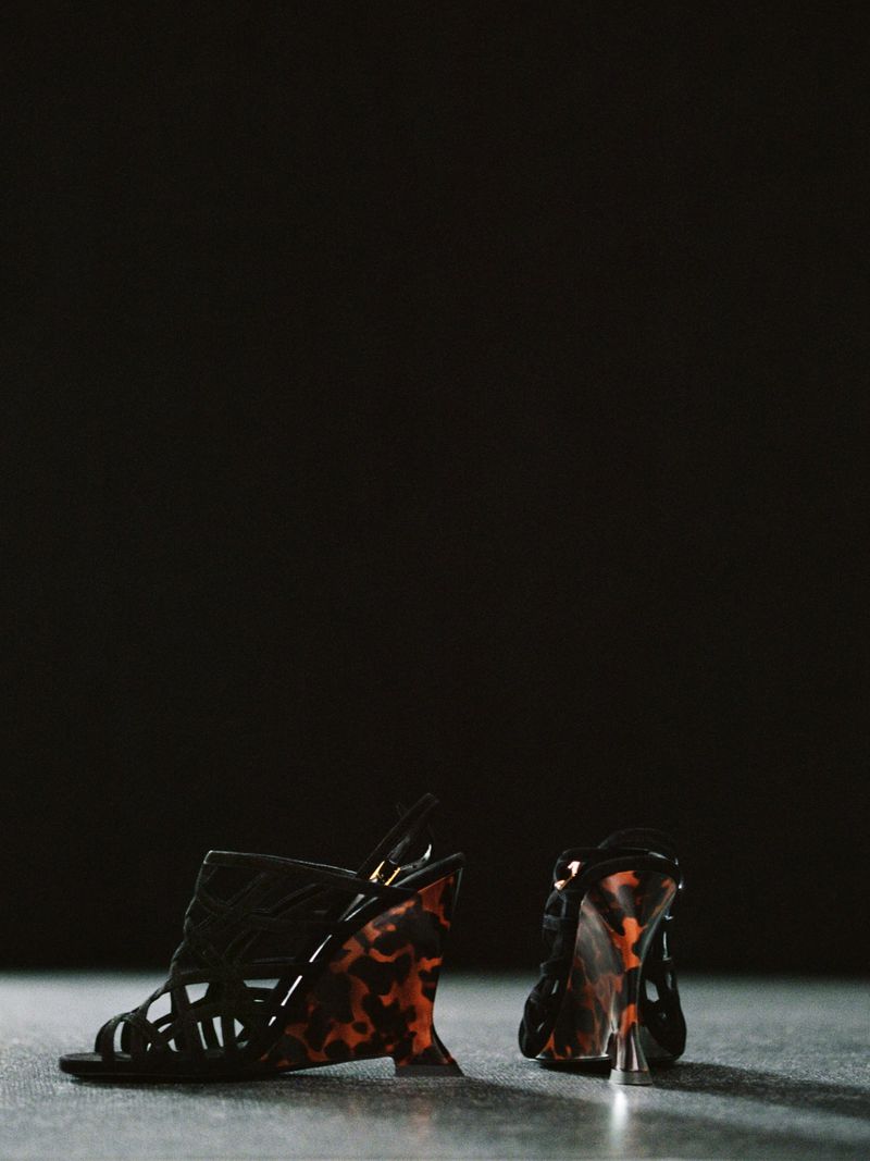 A pair of black cage-style wedge sandals with patterned tortoiseshell-effect heels resting on a dark surface against a black background.