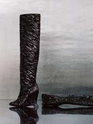 Textured black high-heeled knee boots on a reflective surface against a metallic silver backdrop