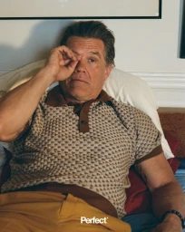 Actor Josh Brolin reclining in bed wearing a patterned knit polo shirt and yellow trousers while rubbing his eye.