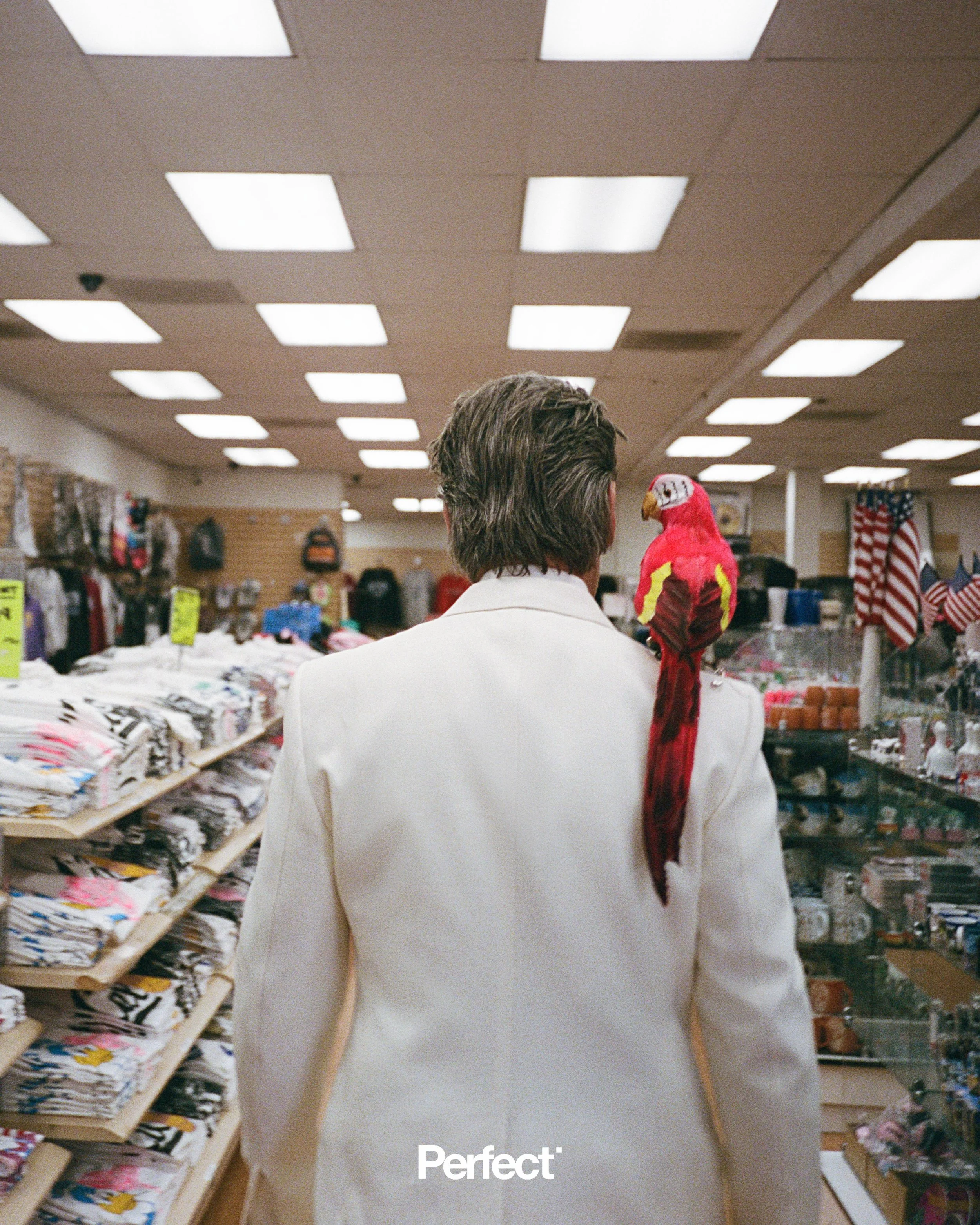 Josh Brolin seen from behind in a white suit jacket with a red parrot perched on his shoulder inside a store aisle.