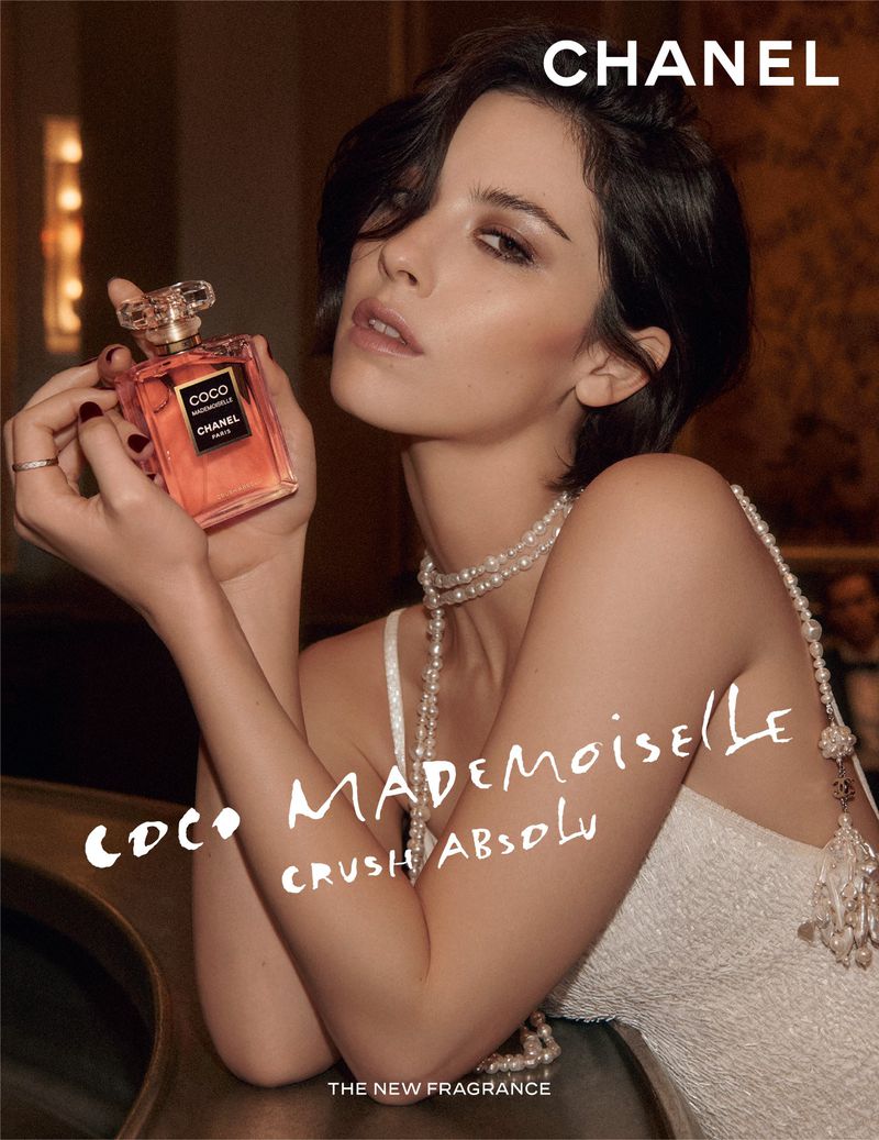 A woman with dark hair holds a bottle of Chanel Coco Mademoiselle perfume, wearing a white textured dress and long pearl necklaces.