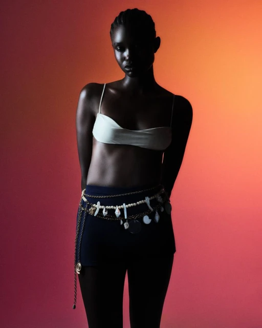 Model poses in a white top and beaded belt against a gradient background. The image captures fashion and beauty in a studio setting.