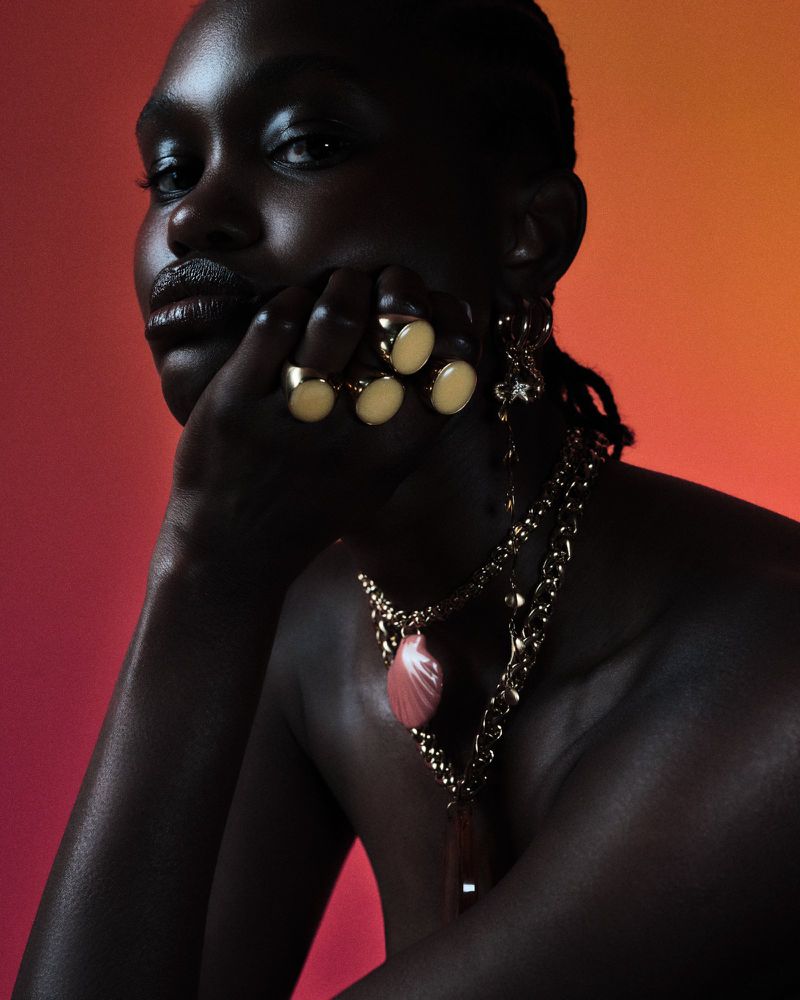 Portrait of a dark-skinned woman wearing gold jewelry, including rings, earrings, and necklaces, against an orange gradient background.