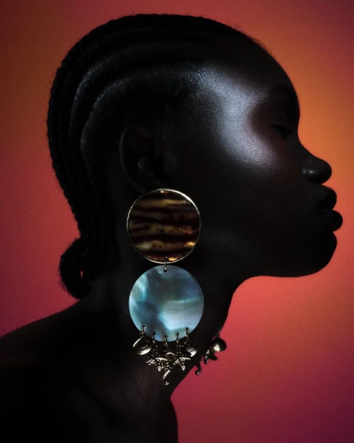 Profile portrait of an adult female with dark skin wearing statement earrings against a graduated red-orange background in studio lighting.