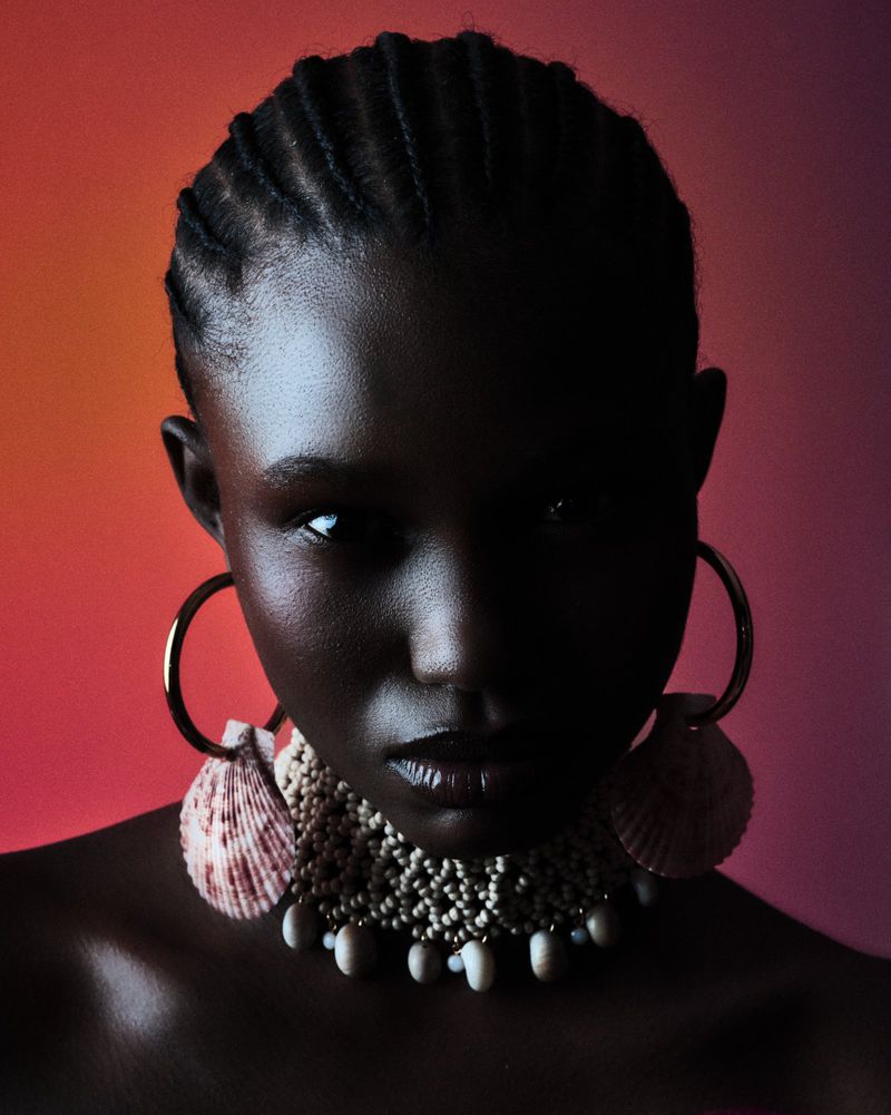 Close-up of a dark-skinned woman with braided hair wearing gold hoop earrings and a shell necklace against a gradient background.
