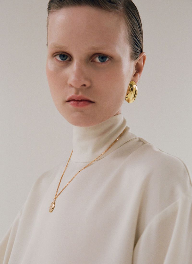 Model Sofia Hansson wearing an ivory turtleneck with gold jewelry and a gold earring against a studio backdrop.
