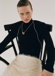 Model Sofia Hansson wearing a black turtleneck and gold pendant necklace seated in a black wooden chair against a white background.