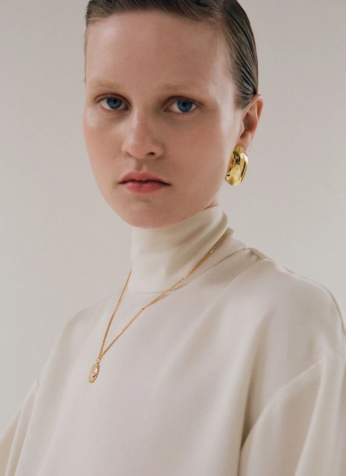Model Sofia Hansson wearing an ivory turtleneck with gold jewelry and a gold earring against a studio backdrop.