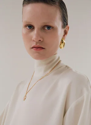 Model Sofia Hansson wearing an ivory turtleneck with gold jewelry and a gold earring against a studio backdrop.