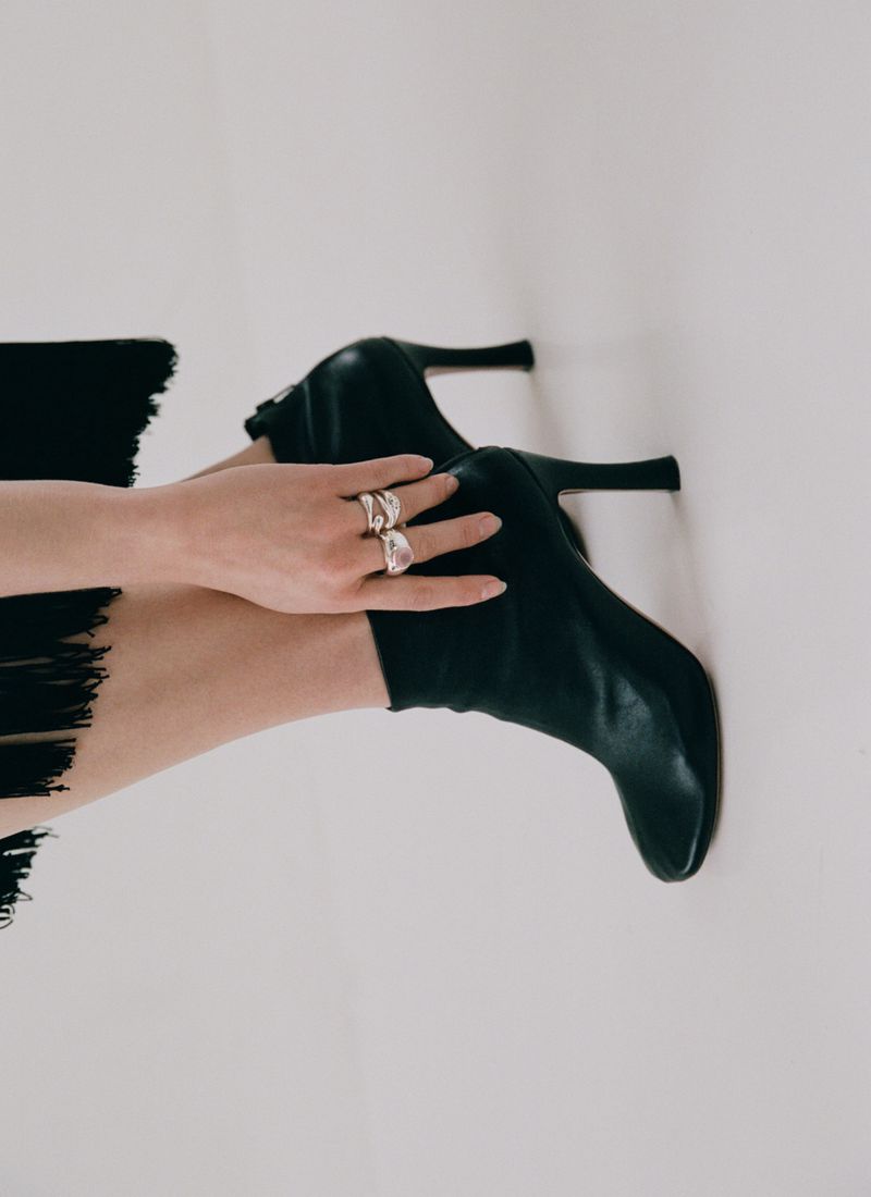 Close-up of a model wearing black heeled ankle boots and sculptural silver rings against a white seamless studio background.