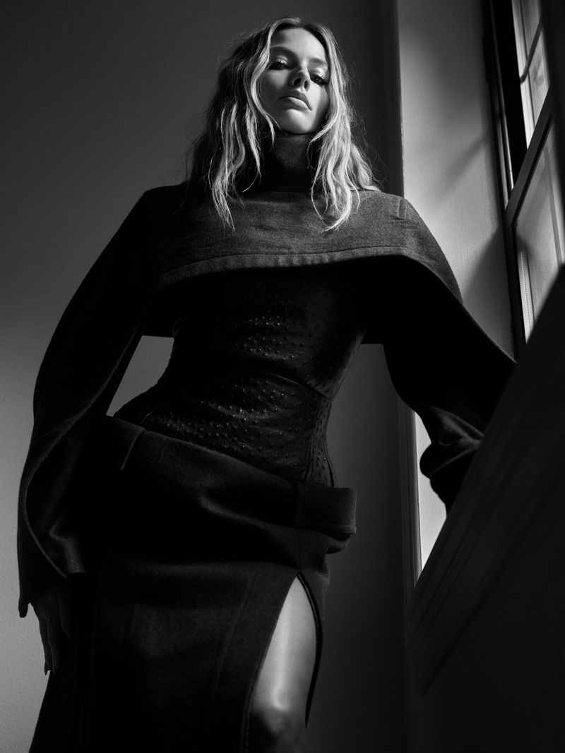 Blonde woman in a dark high-fashion dress, with a high neckline and long sleeves, stands near a window in a moody, low-angle indoor shot.