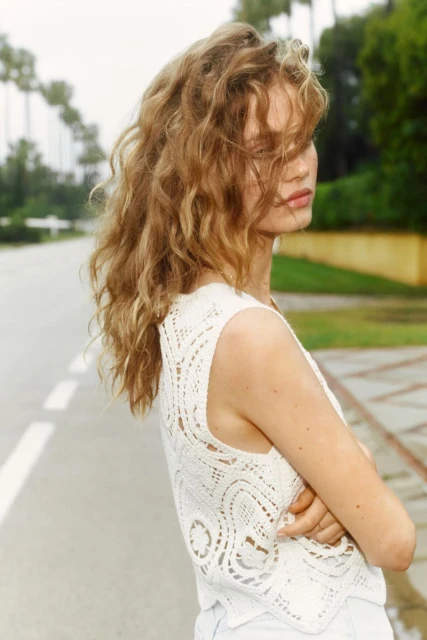 Young woman with curly blonde hair poses in a white crocheted top and light denim outside near green lawns and tall palm trees.