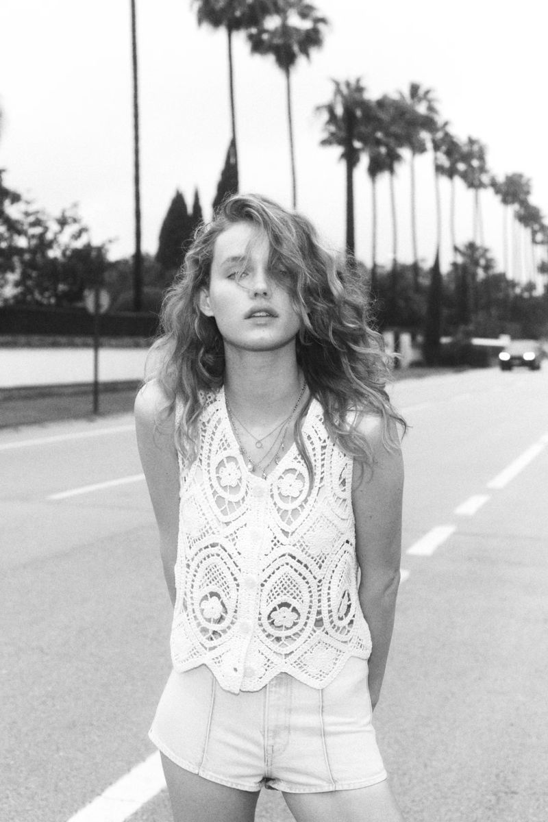Black and white portrait of a young woman with curly hair, wearing a lace top and shorts, standing on a road lined with palm trees.
