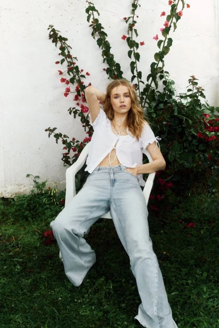A young woman with blonde hair sits on a white chair outdoors, wearing a white blouse and light blue denim pants with vines behind her.