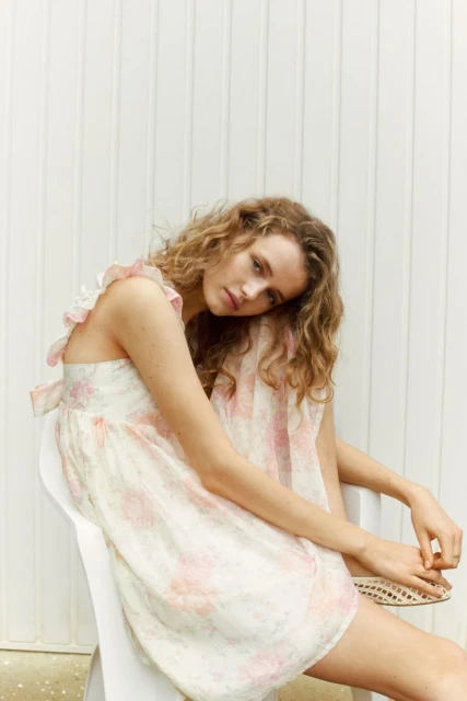 A fair-skinned woman with curly blonde hair sits on a white chair in a floral dress with a white wall as a backdrop.