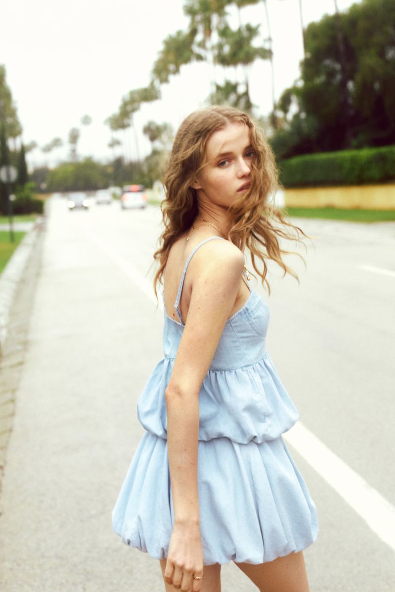 Young woman in a short blue dress stands on a road lined with palm trees on a sunny day, posing for a fashion shot.