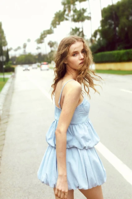 Young woman in a short blue dress stands on a road lined with palm trees on a sunny day, posing for a fashion shot.