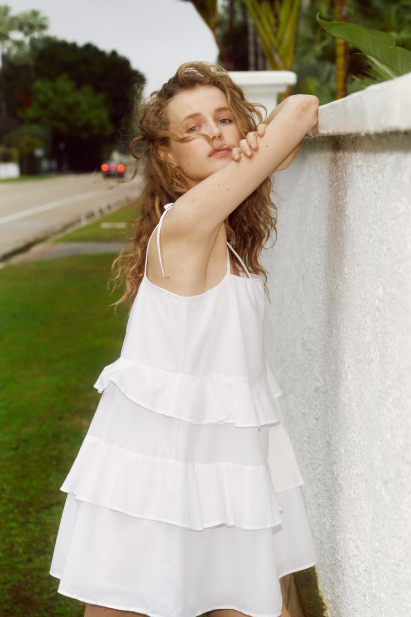 A young adult woman with curly hair poses in a white layered dress against a white concrete wall outdoors with greenery in the background.