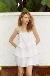 Model wearing a white tiered dress standing against a textured white wall with tropical leaves overhead for an H&M Malaga lookbook.