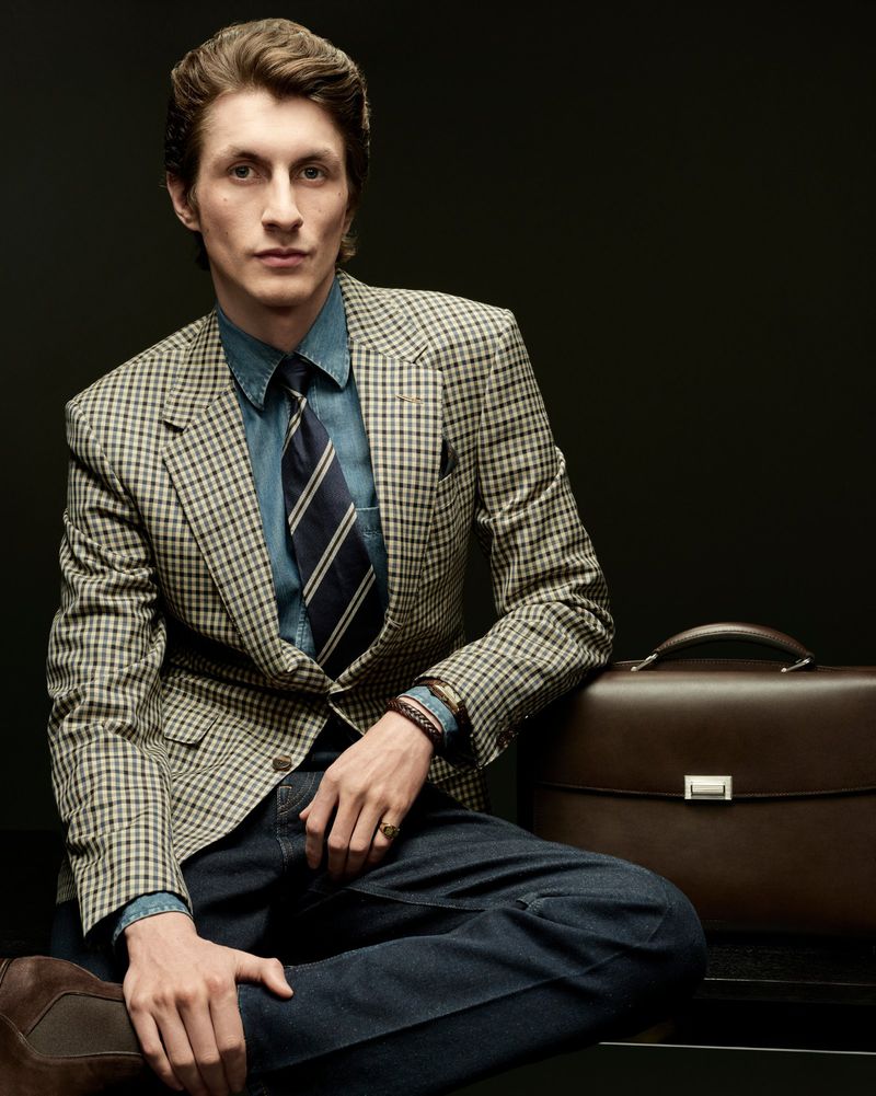 A man in a tan houndstooth blazer and denim shirt sits beside a dark brown leather briefcase against a solid black background.