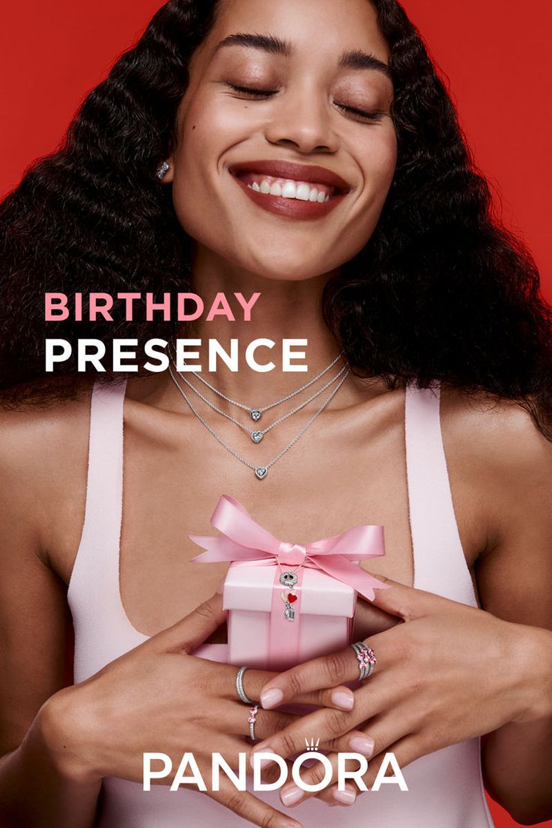 Smiling woman in a red studio setting presents a pink gift box adorned with a bow and Pandora jewelry: necklaces and rings.