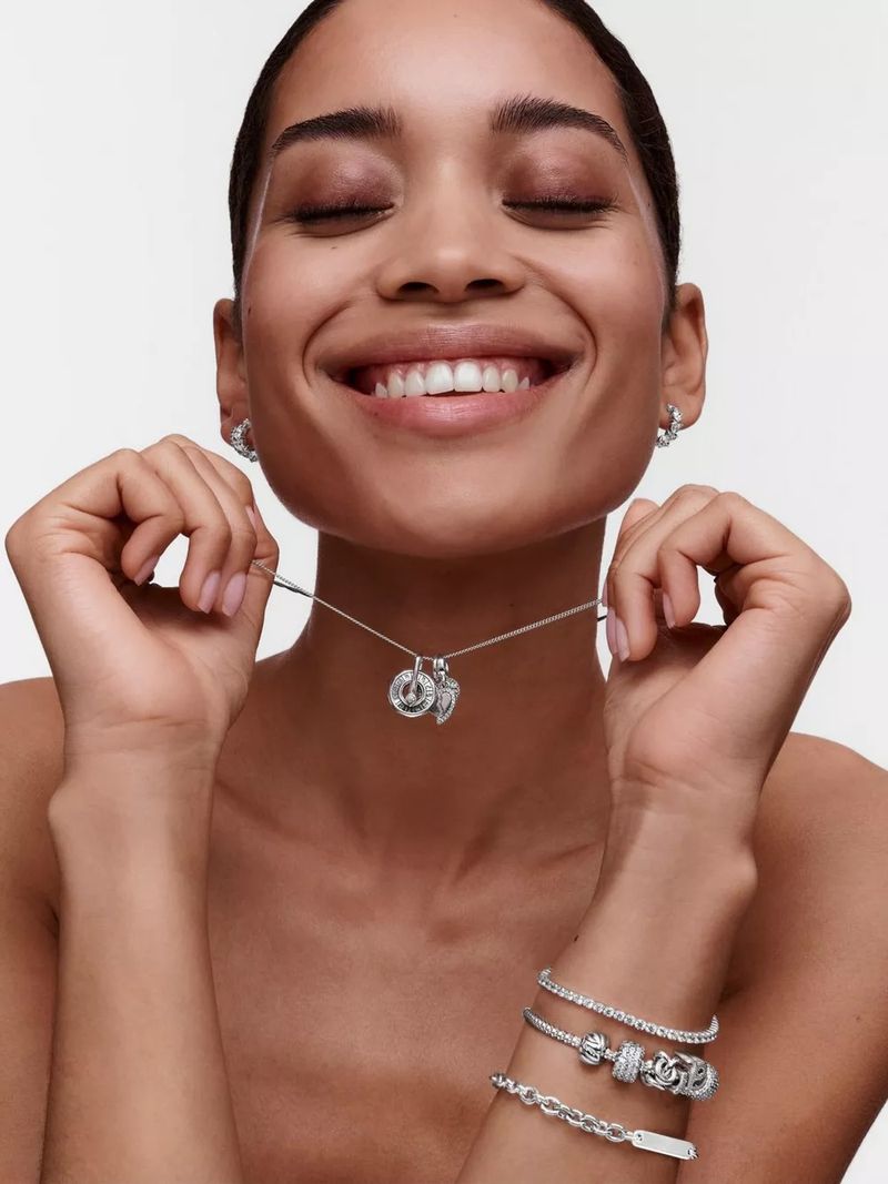 A woman with her eyes closed smiles while showing off silver hoop earrings, a pendant necklace, and bracelet in a bright studio.
