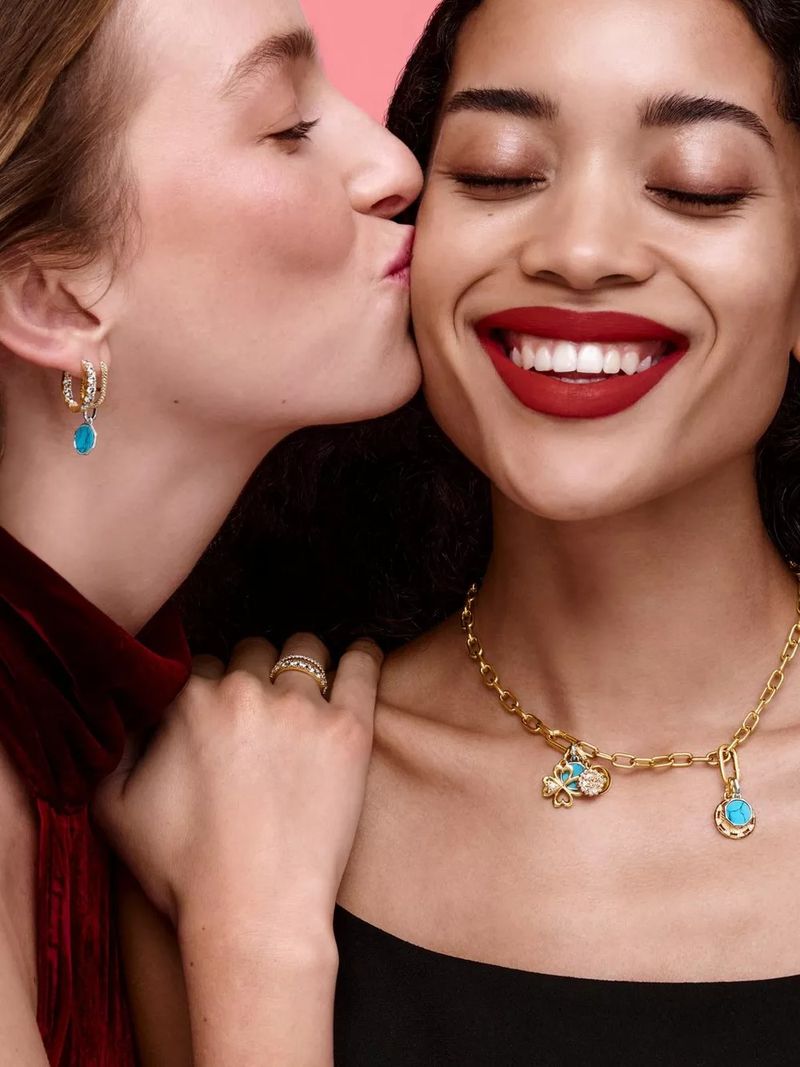 Two women pose in a close-up, one kissing the other's cheek while wearing jewelry against a pink backdrop in studio lighting.