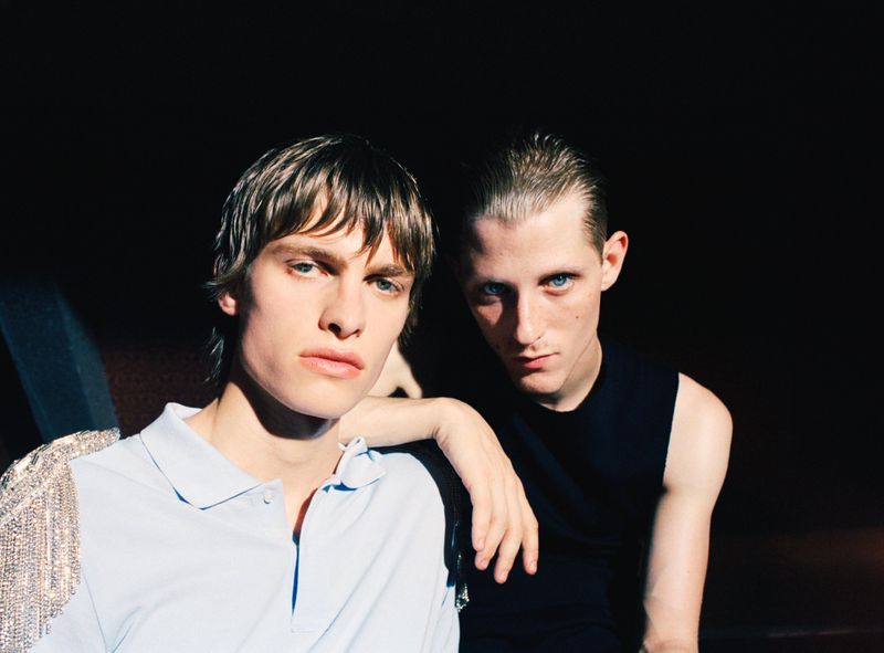 Two male models posing under direct sunlight against a dark background, one in a light blue polo shirt and the other in a black top.