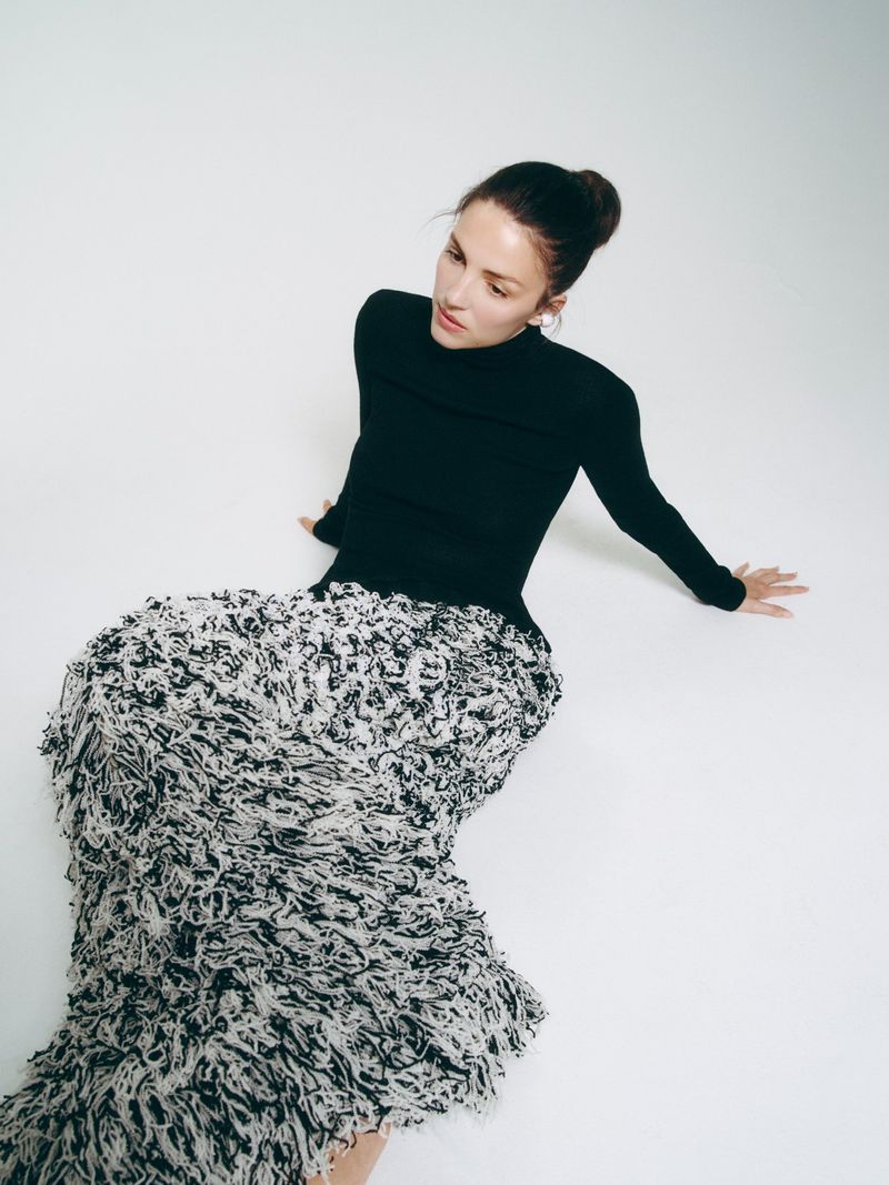 Ella Hunt seated on a white floor wearing a black long-sleeve turtleneck and a voluminous textured black and white fringe skirt.