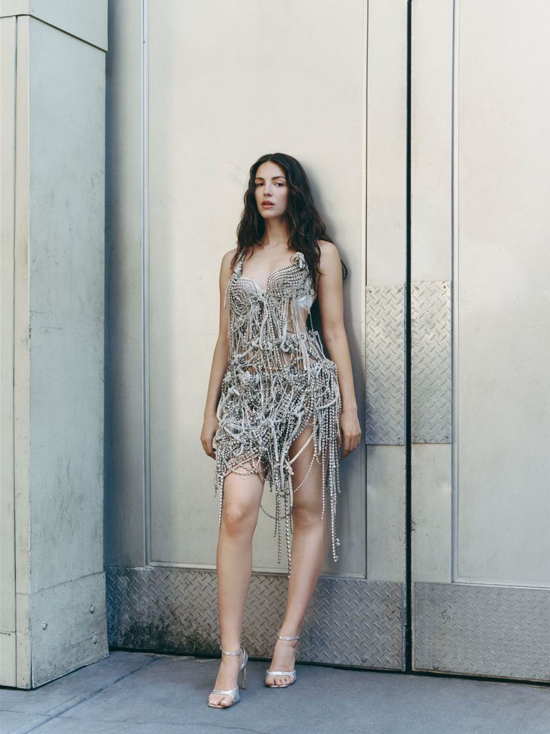 Ella Hunt posing in an intricate silver beaded fringe dress and metallic strappy heels against a metal building exterior.