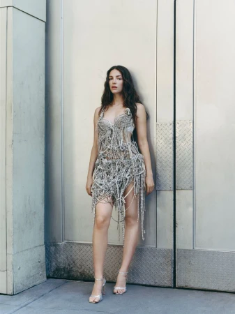 Ella Hunt posing in an intricate silver beaded fringe dress and metallic strappy heels against a metal building exterior.