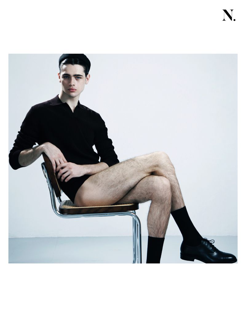 A young man with dark hair wears a black long-sleeved polo and headband, seated on a modern cane-backed chair against a white background.