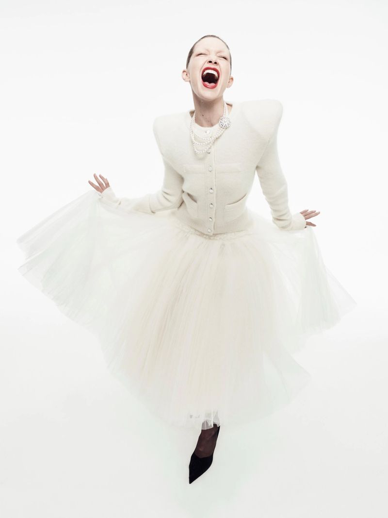 Model with shaved head laughing, wearing a cream knit cardigan with shoulder pads, a white tulle skirt, and black pointed heels.