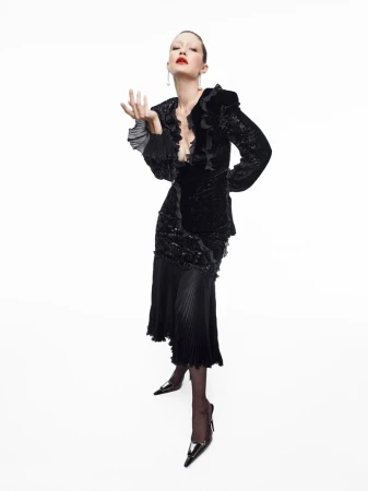 Gigi Hadid posing in a black ruffled blazer and pleated skirt against a white studio background, wearing red lipstick and heels.