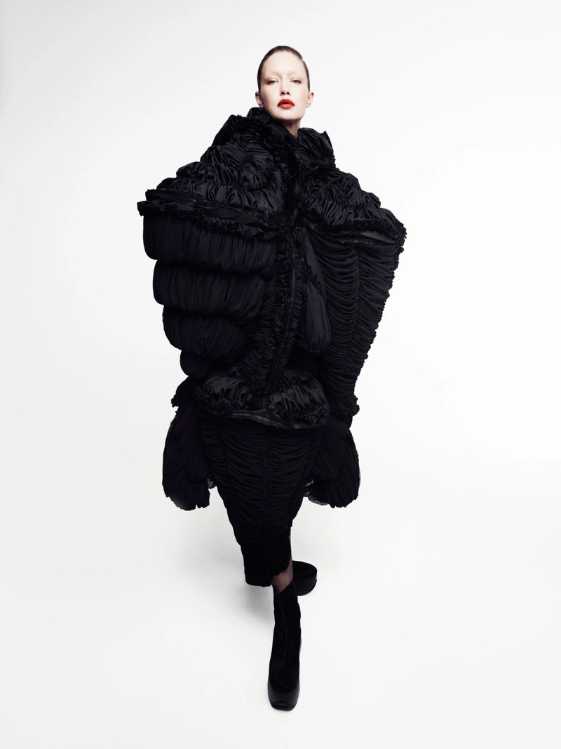 Model with slick hair and red lipstick wears a voluminous, sculptural black pleated coat against a clean white background.