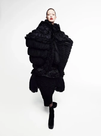 Model with slick hair and red lipstick wears a voluminous, sculptural black pleated coat against a clean white background.
