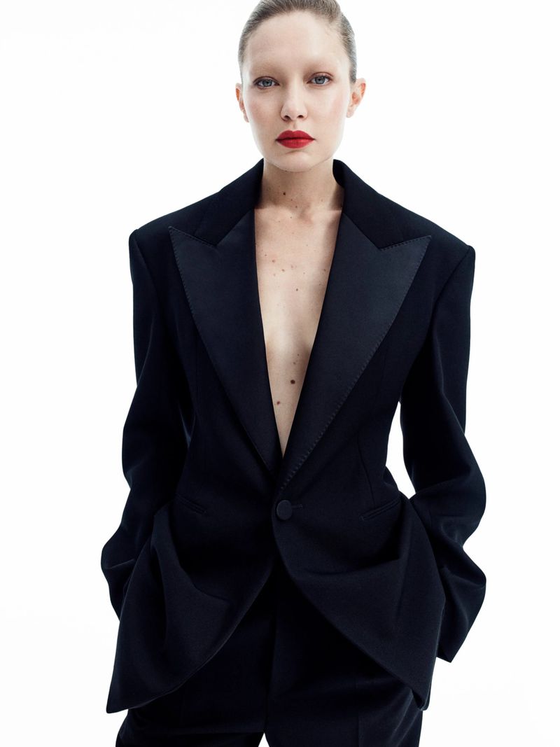 A woman with bleached eyebrows and red lipstick wears a structured black tuxedo jacket with satin lapels against a plain white background.