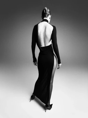 A woman stands with her back to the camera, wearing a long-sleeved black dress with a deep open back and a high side slit.
