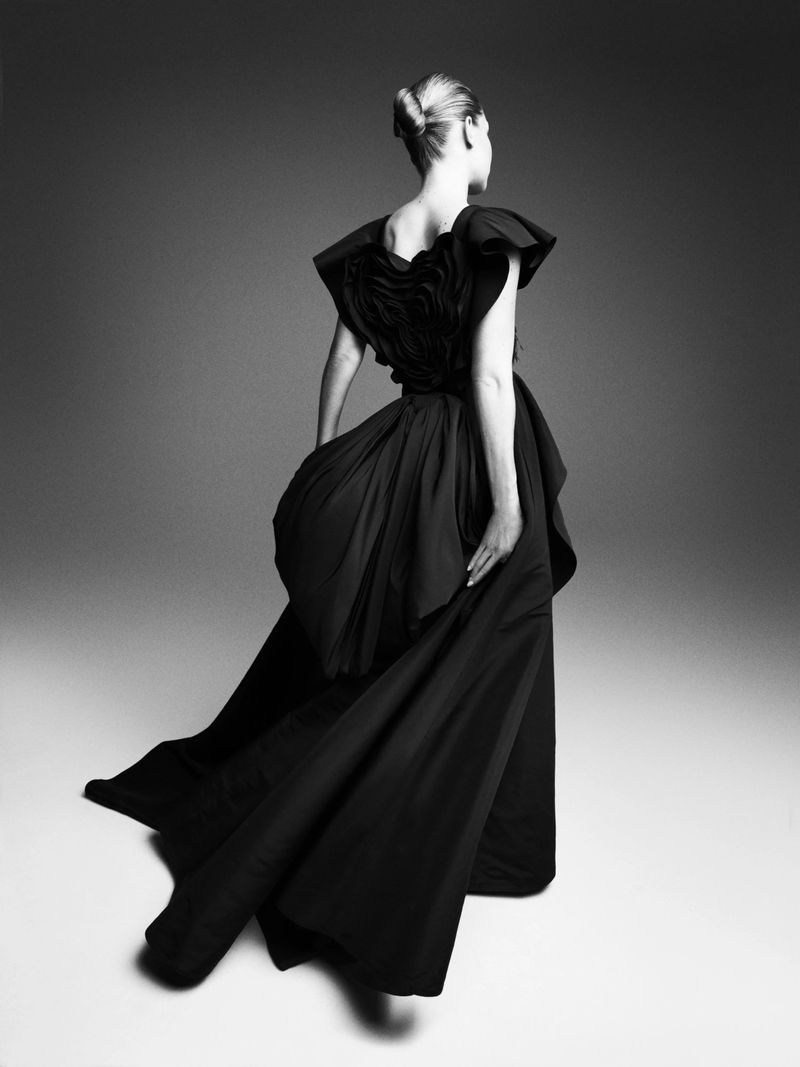 A woman stands with her back to the camera wearing a floor-length black couture dress with ruffled detailing and a full skirt.