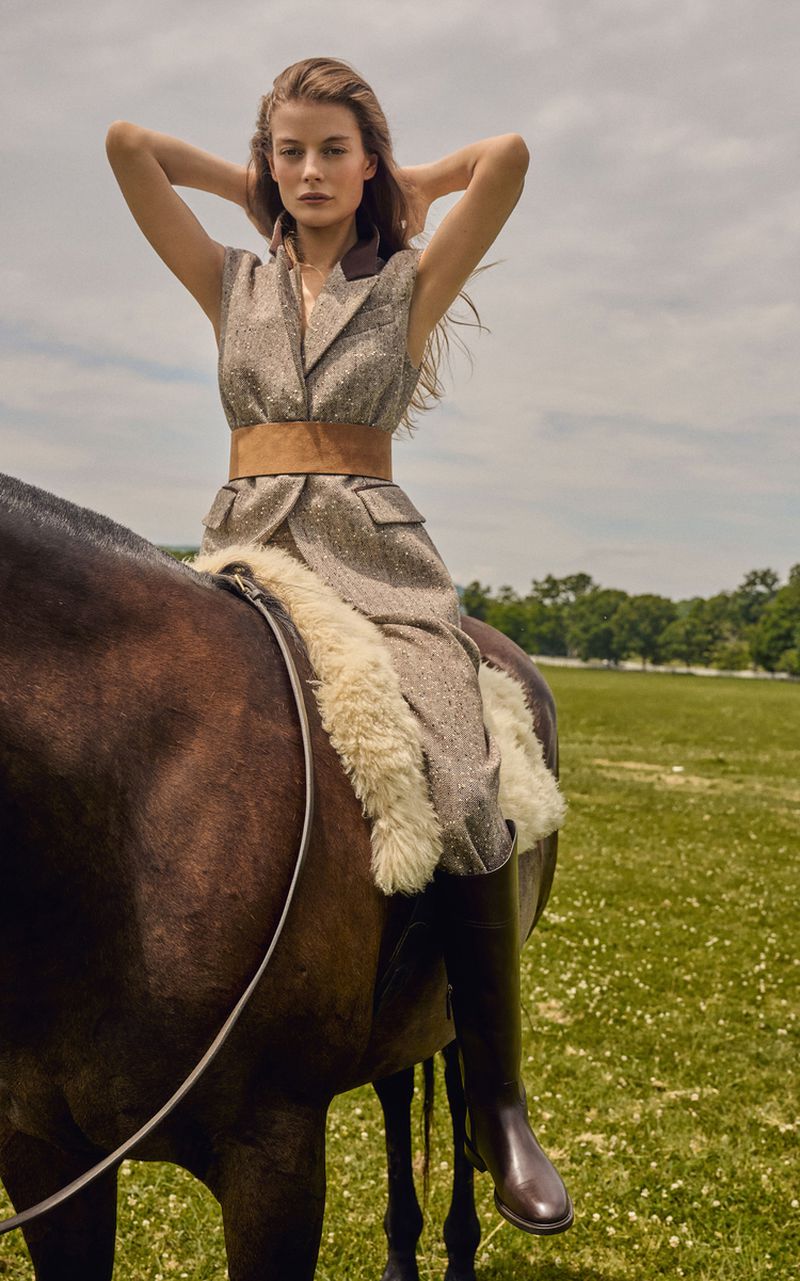 Model Georgia Chambers wearing a tailored tweed sleeveless vest dress and tall boots while riding a horse in a grassy field.