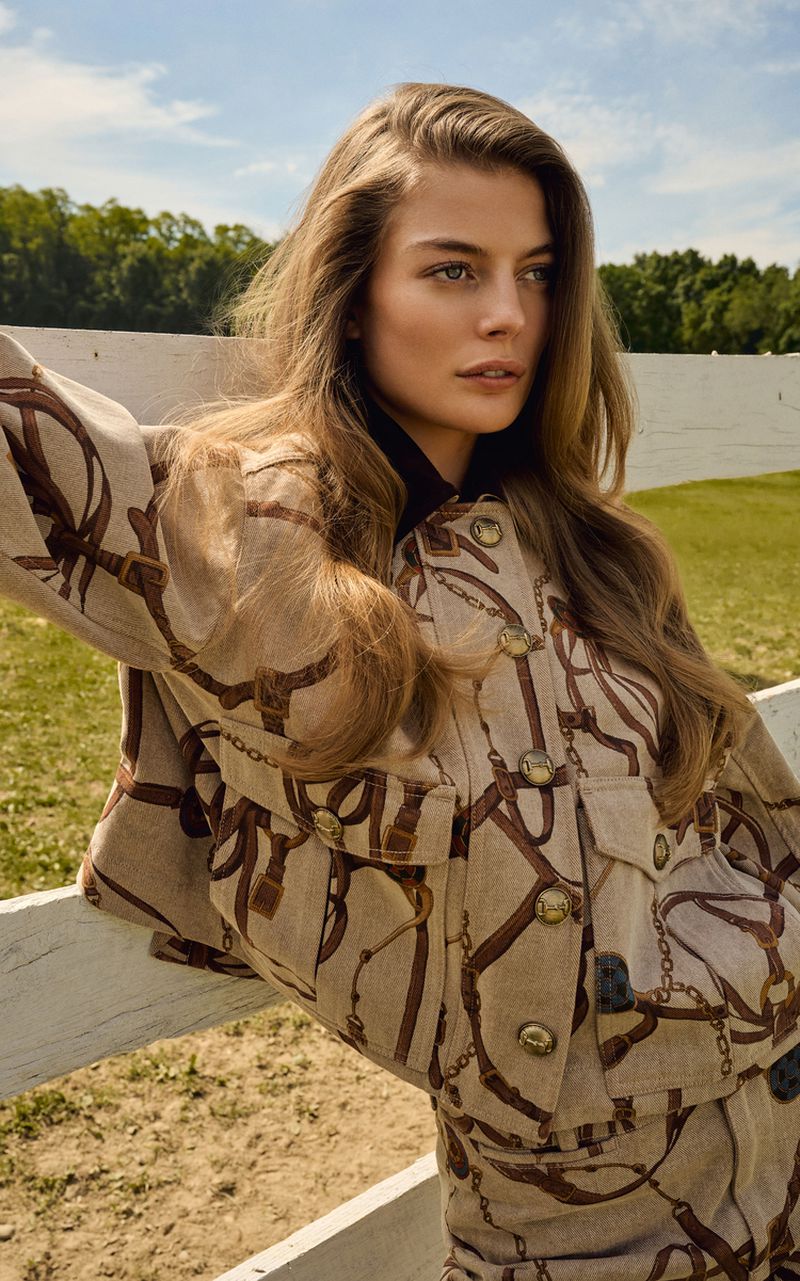 Model Georgia Chambers wearing a beige equestrian harness print jacket while leaning against a white wooden fence outdoors.