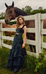 Georgia Chambers posing by a white wooden fence next to a brown horse in a green and blue plaid matching maxi set.