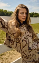 Model Georgia Chambers wearing a beige equestrian harness print jacket while leaning against a white wooden fence outdoors.