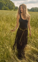 Georgia Chambers posing in a black tank top and brown fringe skirt in a sunlit tall grass field.