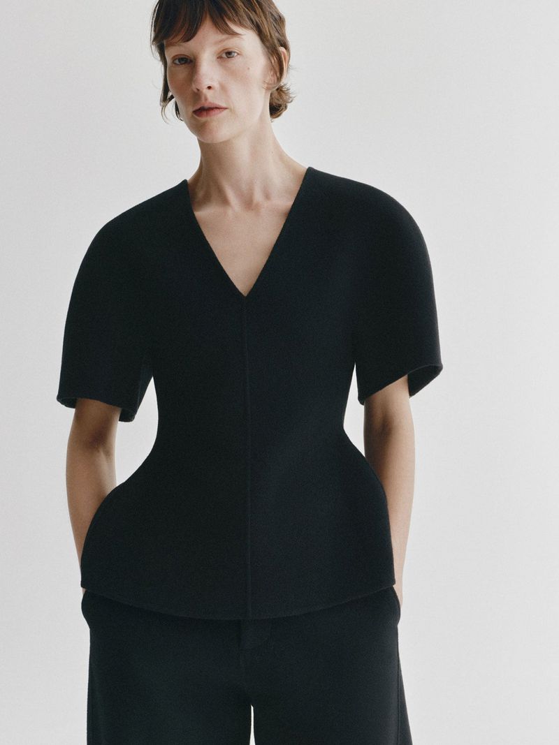 Model Sara Blomqvist wearing a sculpted black V-neck short-sleeve top and matching black trousers against a white seamless background.