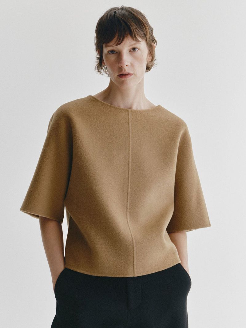 Model Sara Blomqvist wearing a camel short-sleeved knit top and black trousers against a white backdrop.