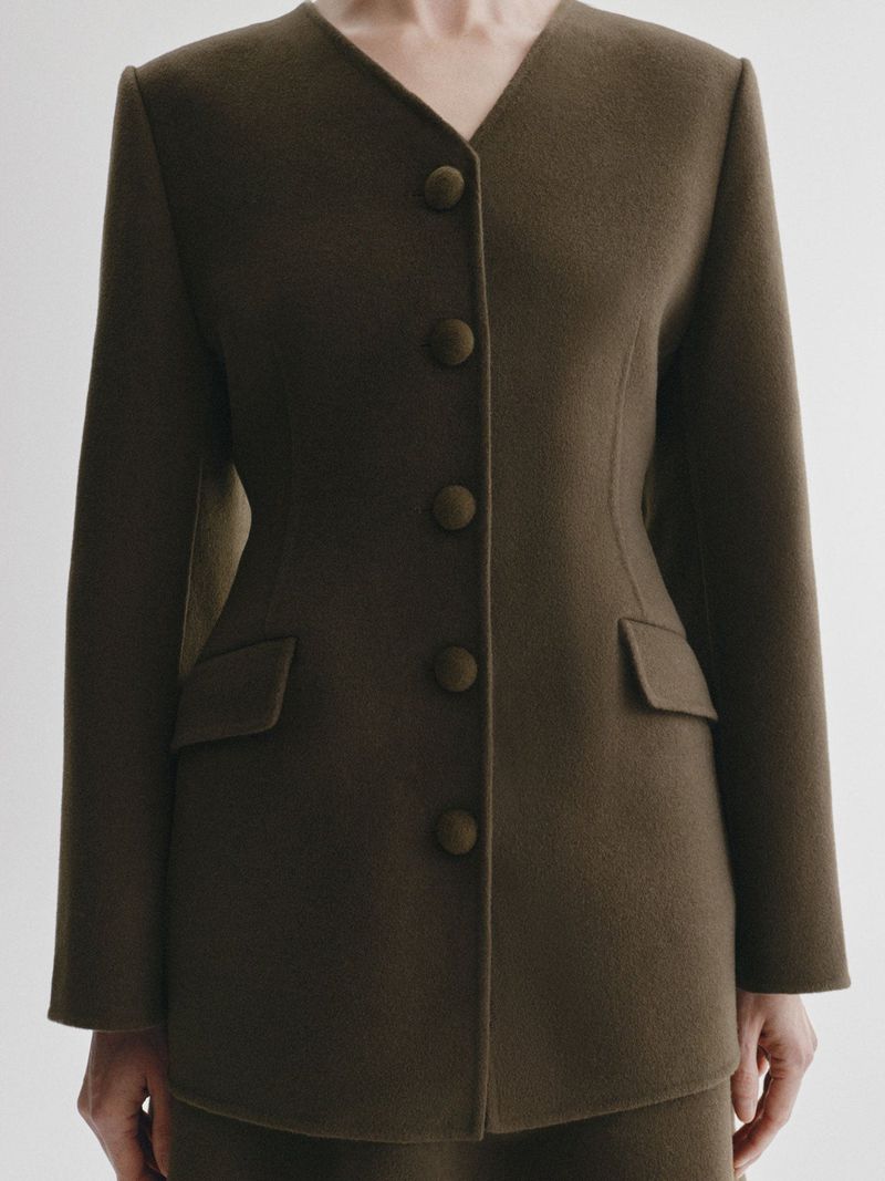 Close-up of a model wearing a tailored collarless olive brown wool buttoned jacket with flap pockets against a white studio background.
