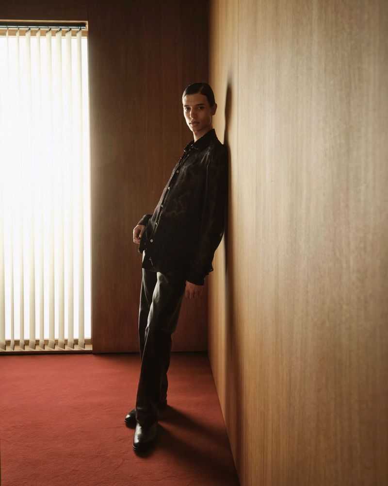 Model Skander Gueye leans against a warm wood-paneled wall wearing an all-black shirt and trousers on red carpet.