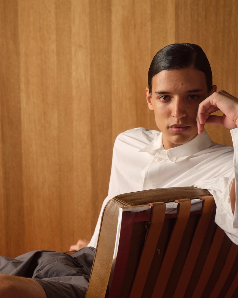 A person with dark hair pulled back wears a crisp white button-down shirt while seated in a modern leather and strap chair.
